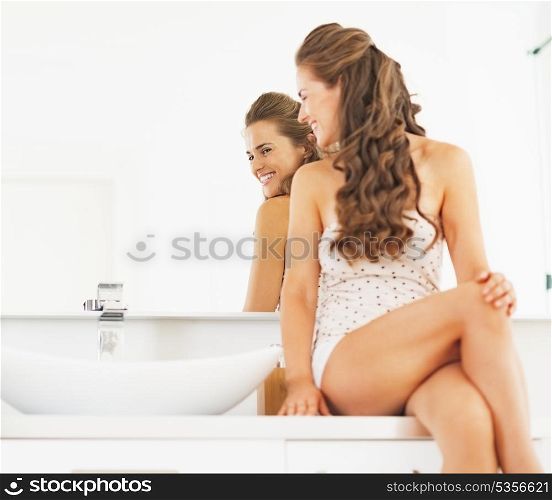 Happy young woman sitting in bathroom and looking in mirror