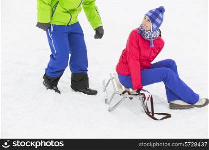 Happy young woman sitting on sled while looking at man in snow