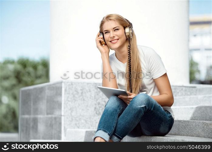 Happy young woman sitting on stairs, listening to the music in music headphones, surfing internet on tablet and buying music online.
