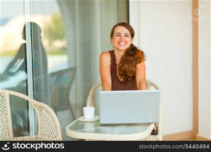 Happy young woman sitting on terrace with cup of coffee and laptop&#xA;
