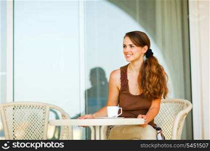 Happy young woman sitting on terrace with cup of tea and looking in corner&#xA;