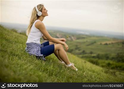Happy young woman sitting outside on grass and listening to music on headphones
