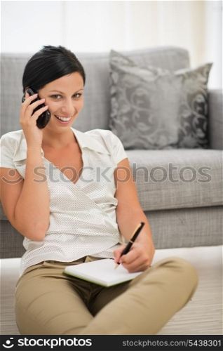 Happy young woman speaking cell phone and writing in notebook
