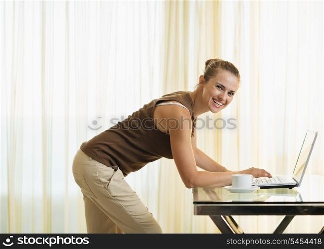 Happy young woman standing near table and working on laptop