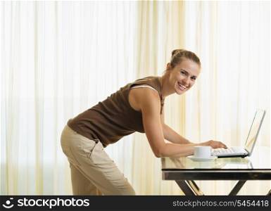 Happy young woman standing near table and working on laptop