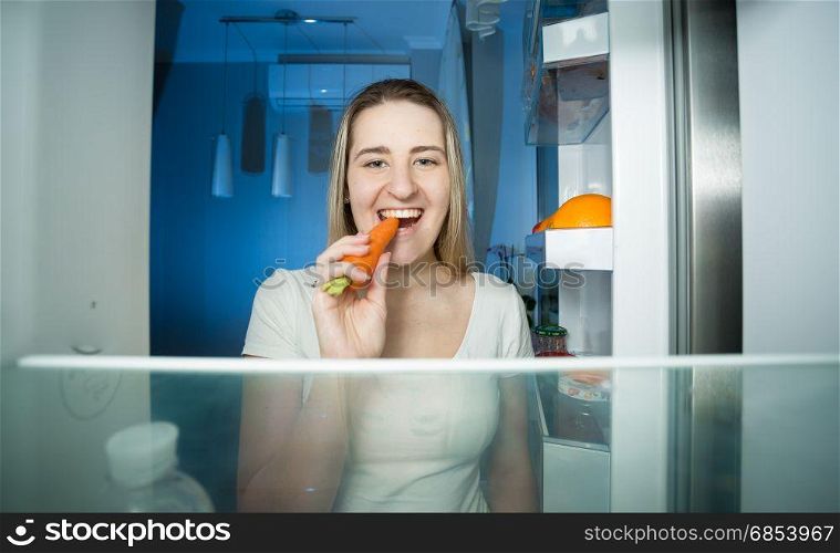 Happy young woman taking carrot from refrigerator and eating it at night