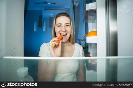 Happy young woman taking carrot from refrigerator and eating it at night