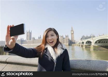 Happy young woman taking self portrait through cell phone against Big Ben at London; England; UK