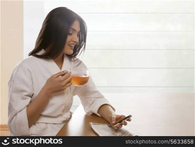 Happy young woman text messaging while holding tea cup at dining table