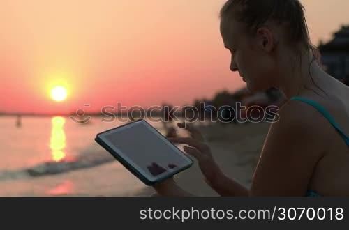 Happy young woman using tablet computer on beach at sunset. Evening sun, sea and people on background