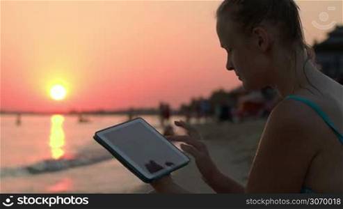 Happy young woman using tablet computer on beach at sunset. Evening sun, sea and people on background
