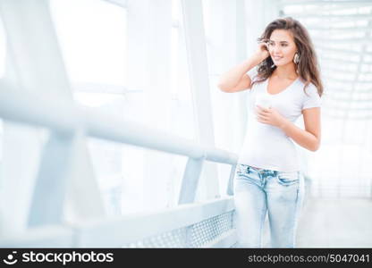 Happy young woman wearing music headphones, standing on the bridge with a take away coffee cup and looking aside against urban background.