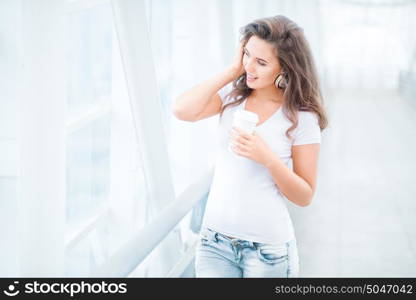 Happy young woman wearing music headphones, standing on the bridge with a take away coffee cup and looking aside against urban background.