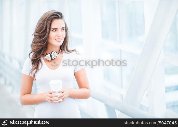 Happy young woman wearing music headphones, standing on the bridge with a take away coffee cup and looking aside against urban background.