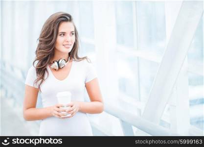 Happy young woman wearing music headphones, standing on the bridge with a take away coffee cup and looking aside against urban background.