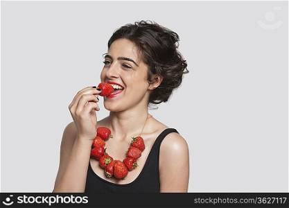 Happy young woman wearing strawberry necklace as she eats one piece over gray background