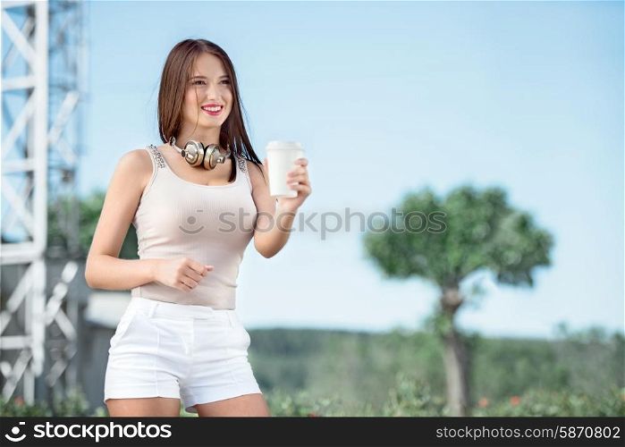 Happy young woman wearing vintage music headphones around her neck, holding takeaway coffee and posing against background of parabolic satellite dishes on towers.