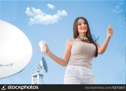 Happy young woman wearing vintage music headphones around her neck, holding takeaway coffee and posing against background of parabolic satellite dishes that receive wireless signals from satellites.