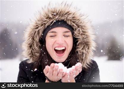 Happy young woman wearing winter clothes while blowing snow on snowy day with snowflakes around her in beautiful winter forest