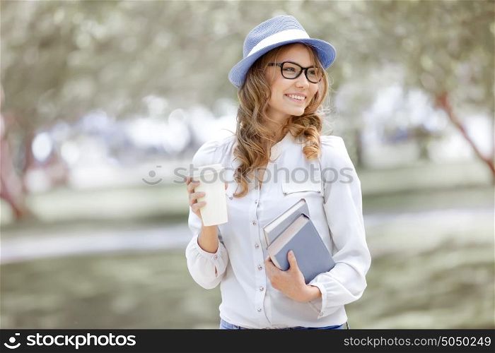 Happy young woman with a coffee-to-go, walking in a summer park and holding books for reading.