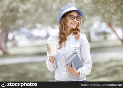 Happy young woman with a coffee-to-go, walking in a summer park and holding books for reading.