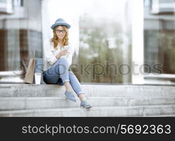 Happy young woman with a disposable coffee cup and shopping bags sitting on the stairs and using her smartphone for communication via wi-fi internet in a summer park.