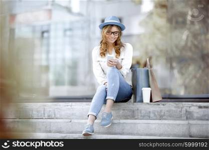 Happy young woman with a disposable coffee cup and shopping bags sitting on the stairs and using her smartphone for communication via wi-fi internet in a summer park.