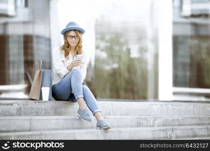 Happy young woman with a disposable coffee cup and shopping bags sitting on the stairs and using her smartphone for communication via wi-fi internet in a summer park.