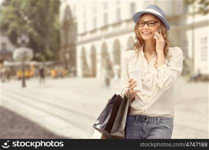 Happy young woman with a disposable coffee cup and shopping bags, talking on phone and smiling against urban city background.