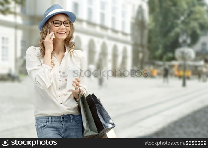 Happy young woman with a disposable coffee cup and shopping bags, talking on phone and smiling against urban city background.