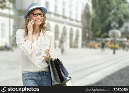 Happy young woman with a disposable coffee cup and shopping bags, talking on phone and smiling against urban city background.