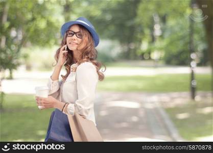 Happy young woman with a disposable coffee cup and shopping bags, talking on phone and smiling against city park background.