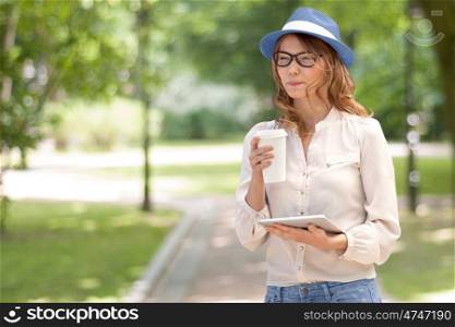 Happy young woman with a disposable coffee cup, holding tablet in her hands, drinking coffee and smiling against summer park background.