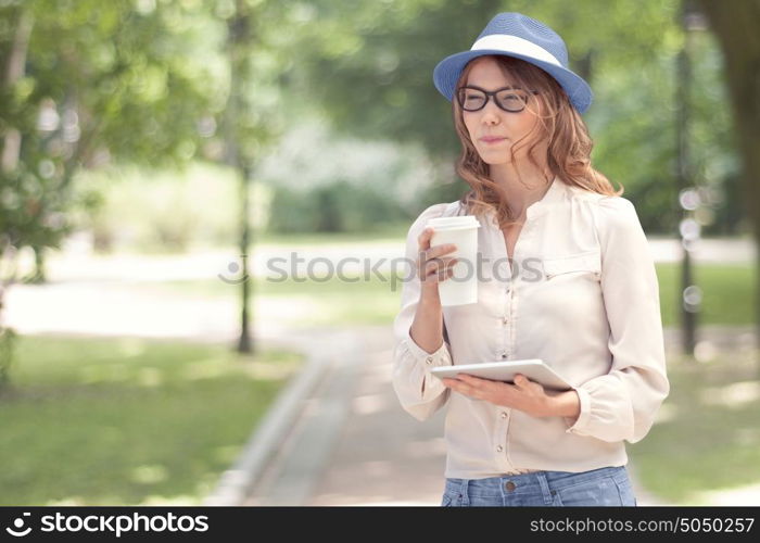 Happy young woman with a disposable coffee cup, holding tablet in her hands, drinking coffee and smiling against summer park background.