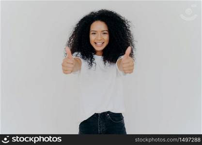 Happy young woman with Afro hairstyle, shows thumbs up, accepts or agrees awesome plan, gives her approval, smiles broadly, stands indoor, says excellent decision, isolated over white background