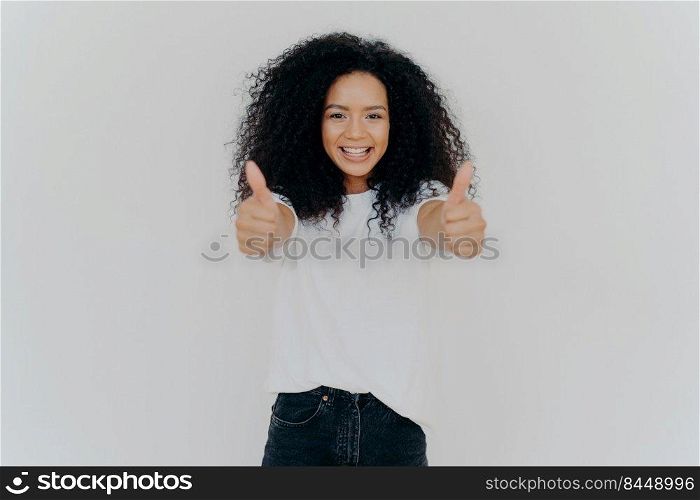 Happy young woman with Afro hairstyle, shows thumbs up, accepts or agrees awesome plan, gives her approval, smiles broadly, stands indoor, says excellent decision, isolated over white background