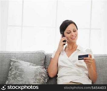 Happy young woman with credit card sitting on sofa and talking mobile phone