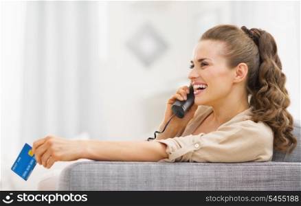 Happy young woman with credit card talking phone in living room