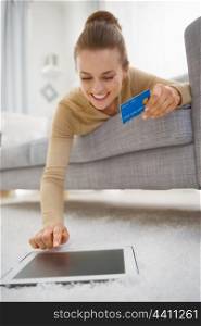 Happy young woman with credit card using tablet pc while laying on sofa