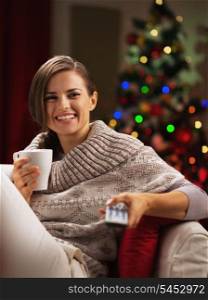 Happy young woman with cup of hot beverage near christmas tree watching tv