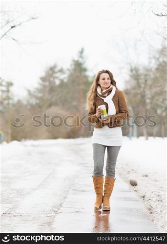 Happy young woman with cup of hot beverage walking in winter park