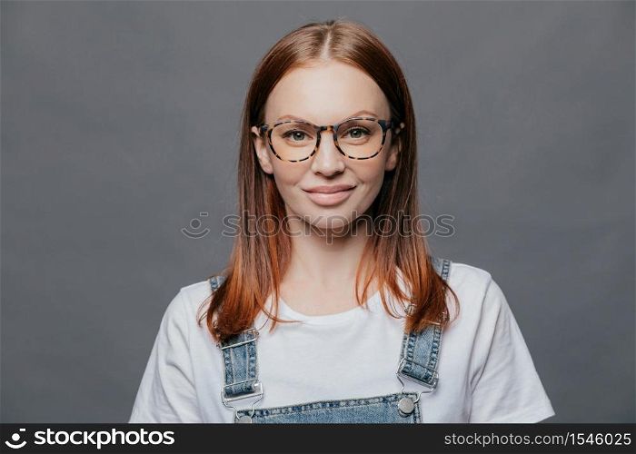 Happy young woman with glad facial expression, has healthy skin, wears optical glasses, white t shirt and denim sarafan, models over grey background, going to meet with friends, poses indoor