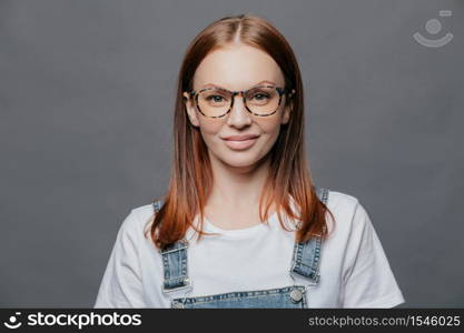 Happy young woman with glad facial expression, has healthy skin, wears optical glasses, white t shirt and denim sarafan, models over grey background, going to meet with friends, poses indoor