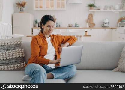 Happy young woman with laptop is sitting on couch at home. Spanish girl is chatting and browsing internet. Remote work or study from home. Cosy living room and kitchen interior.. Happy young woman with laptop is sitting on couch at home. Remote work or study from home.