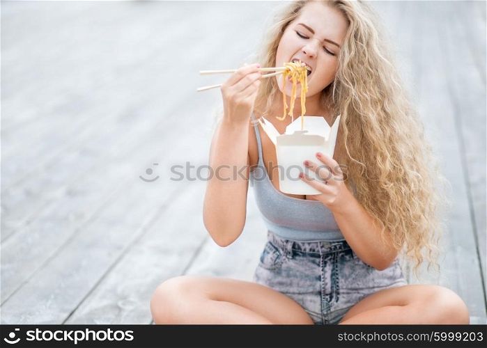 Happy young woman with long curly hair, holding a lunch box and eating up noodles from Chinese take-out with chopsticks.