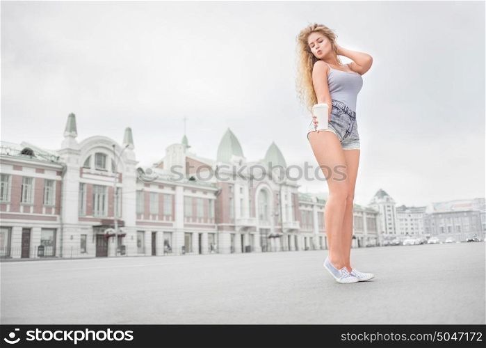 Happy young woman with long curly hair, holding a take away coffee cup and drinking coffee with flirt against urban city background.