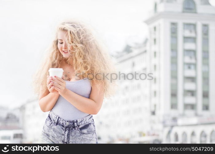 Happy young woman with long curly hair, holding a take away coffee cup and smiling with flirt in front of a camera against urban city traffic background.