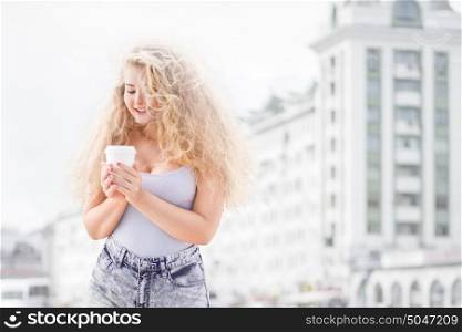 Happy young woman with long curly hair, holding a take away coffee cup and smiling with flirt in front of a camera against urban city traffic background.
