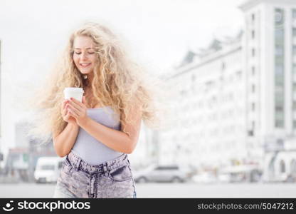 Happy young woman with long curly hair, holding a take away coffee cup and smiling with flirt in front of a camera against urban city traffic background.