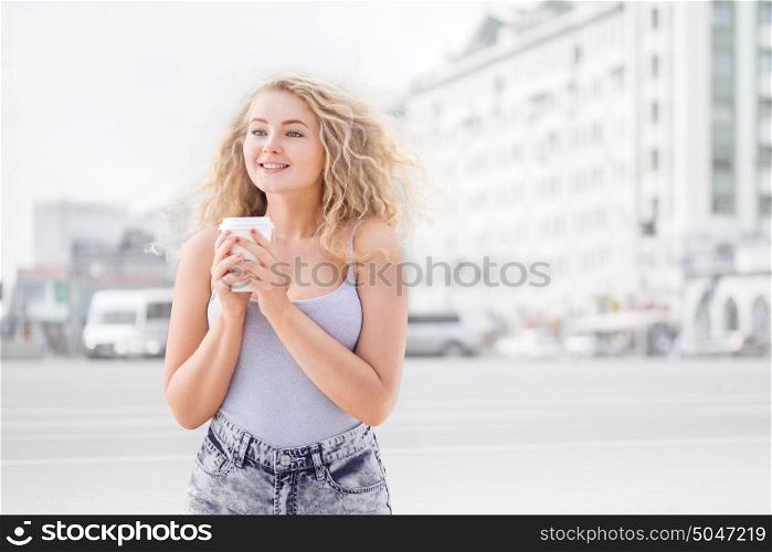 Happy young woman with long curly hair, holding a take away coffee cup and smiling with flirt in front of a camera against urban city traffic background.